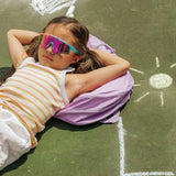 Girl relaxing outdoors wearing Cotton Candy youth sunglasses with UV400 shield lens and pastel-color frame, chalk drawings around her.
