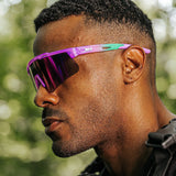 Lifestyle view of LosReyes Prime Jurassic Brontosaurus sunglasses in purple and green, featuring a mirrored shield lens and wraparound athletic frame, worn outdoors for high-performance training and UV400 protection.