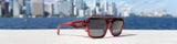 Ares sunglasses from the LosReyes Lifestyle Collection displayed on a sandy surface with a waterfront city skyline behind them.