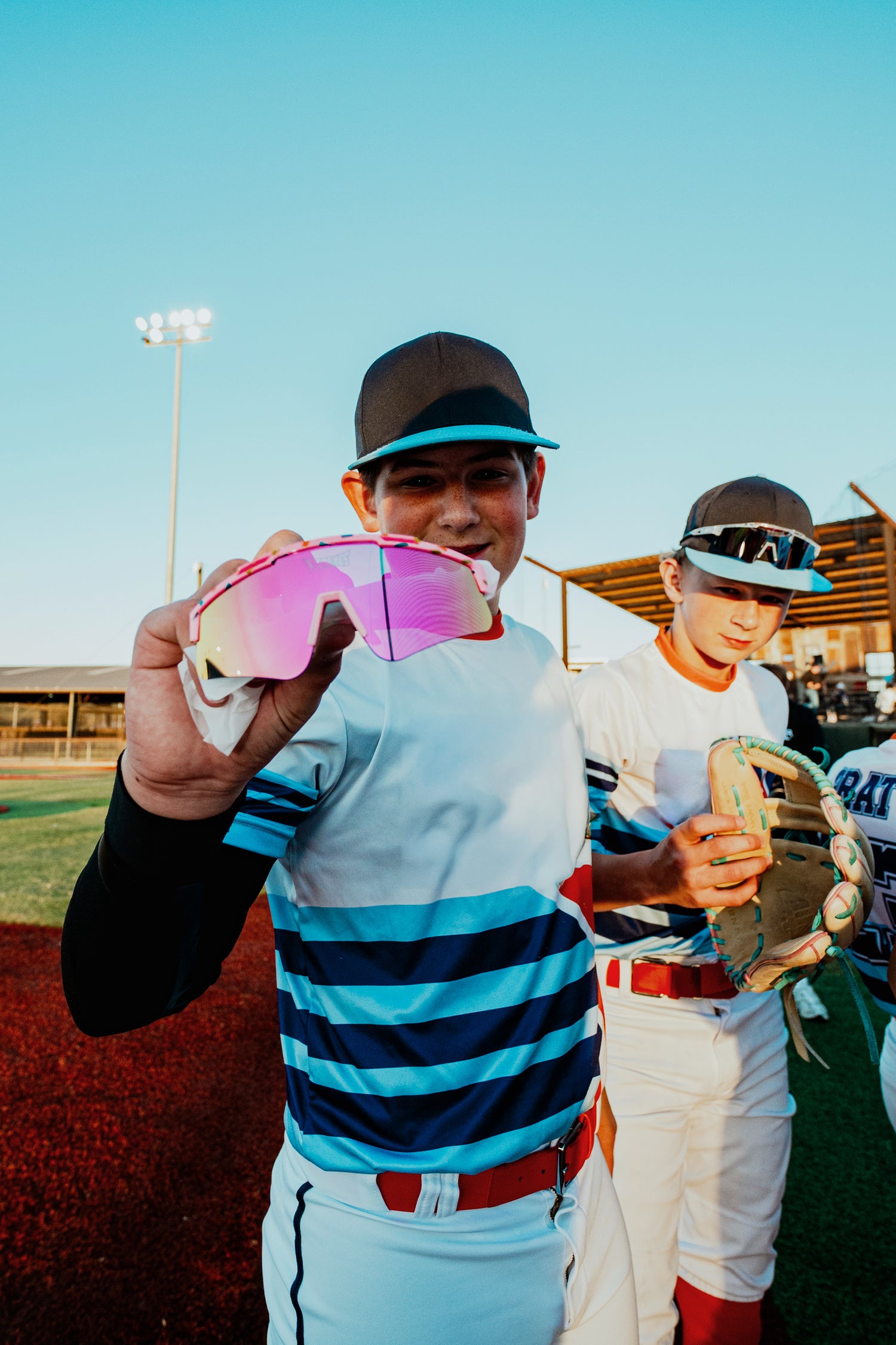 Youth baseball player holding LosReyes Apparel Candy Collection pink mirror sunglasses on baseball field, community sports culture brand