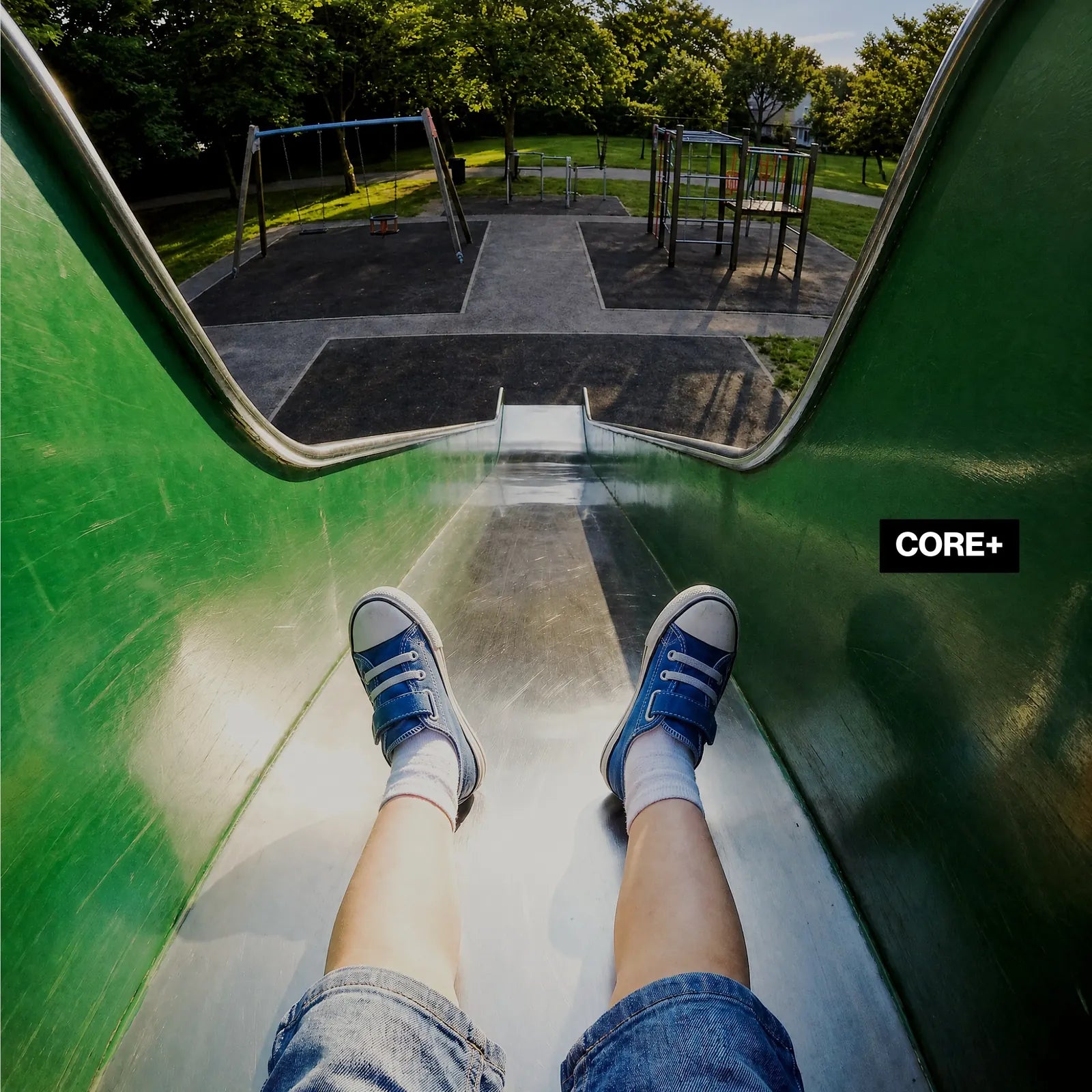 First-person POV looking down a stainless steel playground slide with green sides, wearing blue low-top sneakers and denim shorts, CORE+ campaign badge, park with swings and climbing frames in the background.