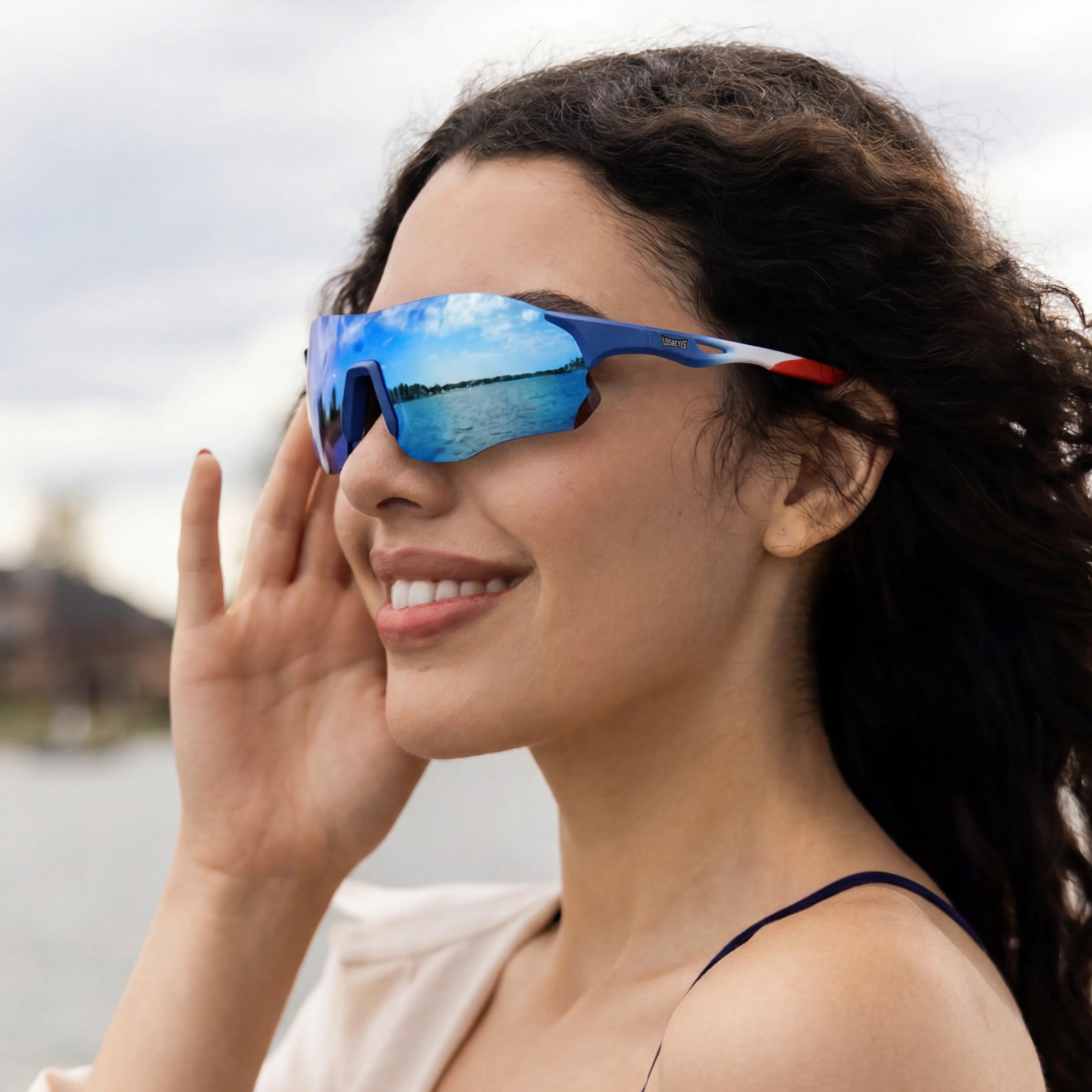 Woman with curly dark hair smiling by the water wearing LOSREYES Edge USA 250 limited edition running sunglasses with red white and blue frame and blue mirrored lens