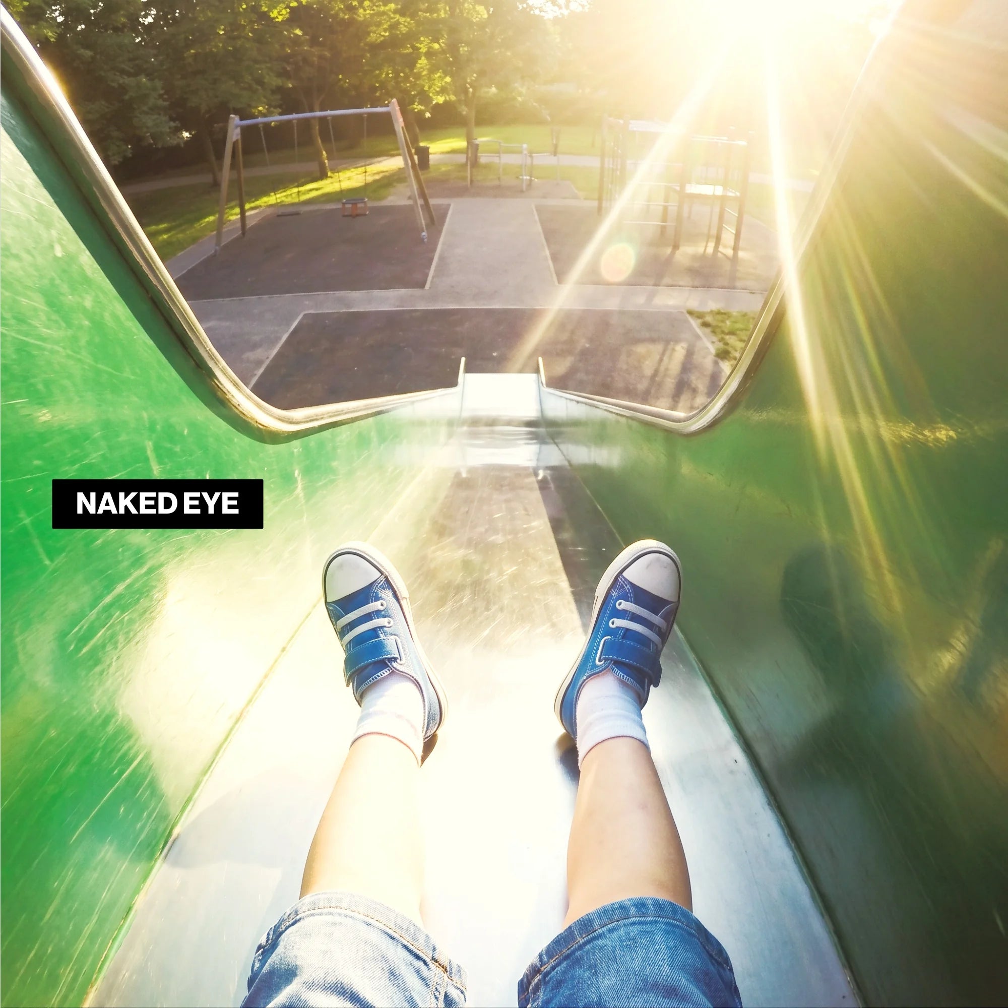 First-person POV down a green and steel playground slide with warm sun flare, blue low-top sneakers and denim shorts, NAKED EYE campaign badge, tree-lined park playground behind.