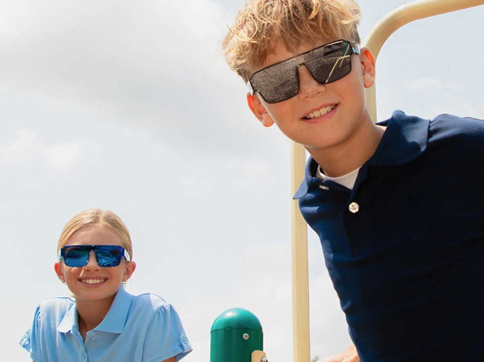 Kids wearing LosReyes Omega youth shield sunglasses outdoors, boy in Hades black reflective lens and girl in Superhero blue mirrored lens, UV400 performance eyewear on a sunny playground