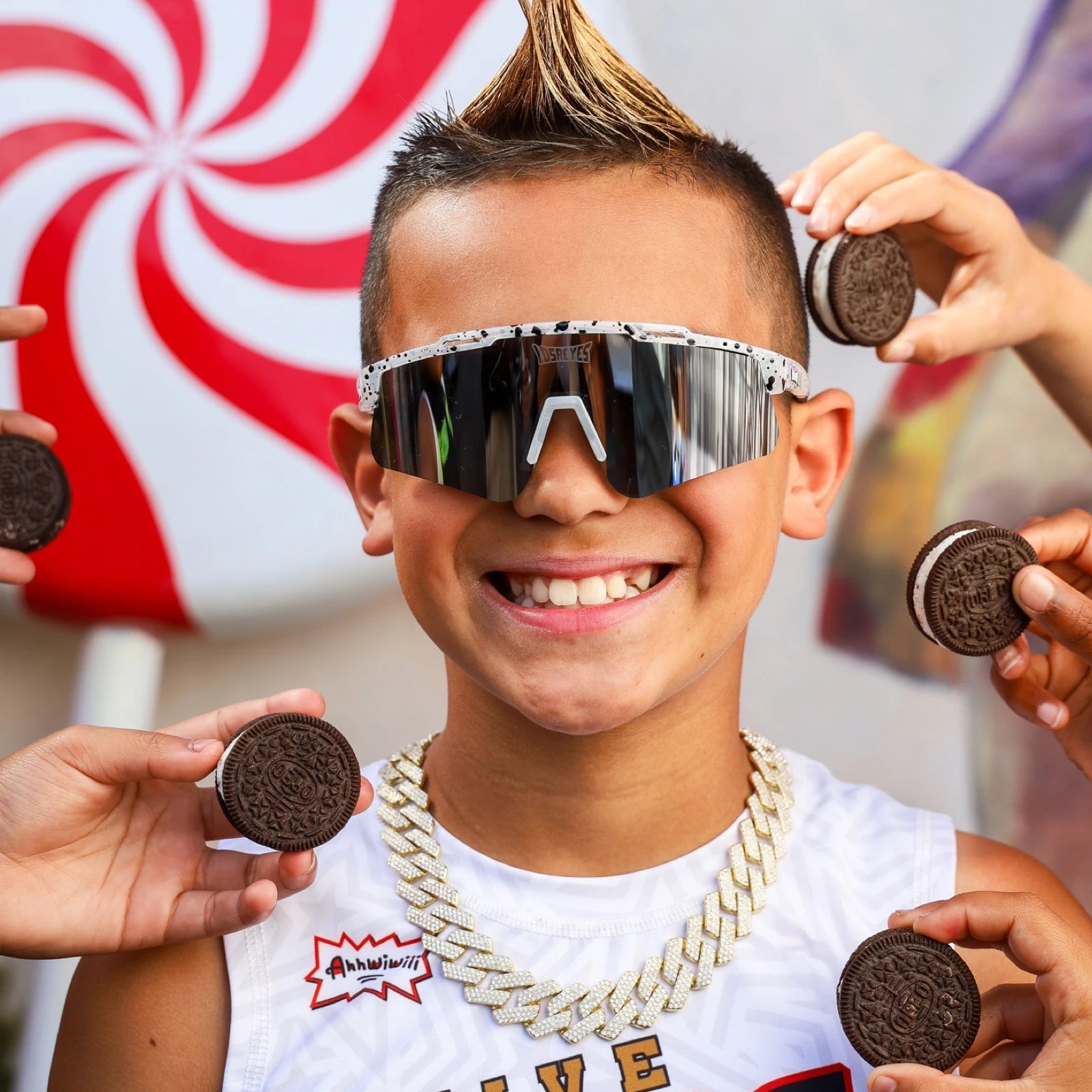 Kid wearing LosReyes Prime Youth Cookie sports sunglasses holding sandwich cookies, showing off mirrored lenses and youth-sized black speckle wraparound frame.