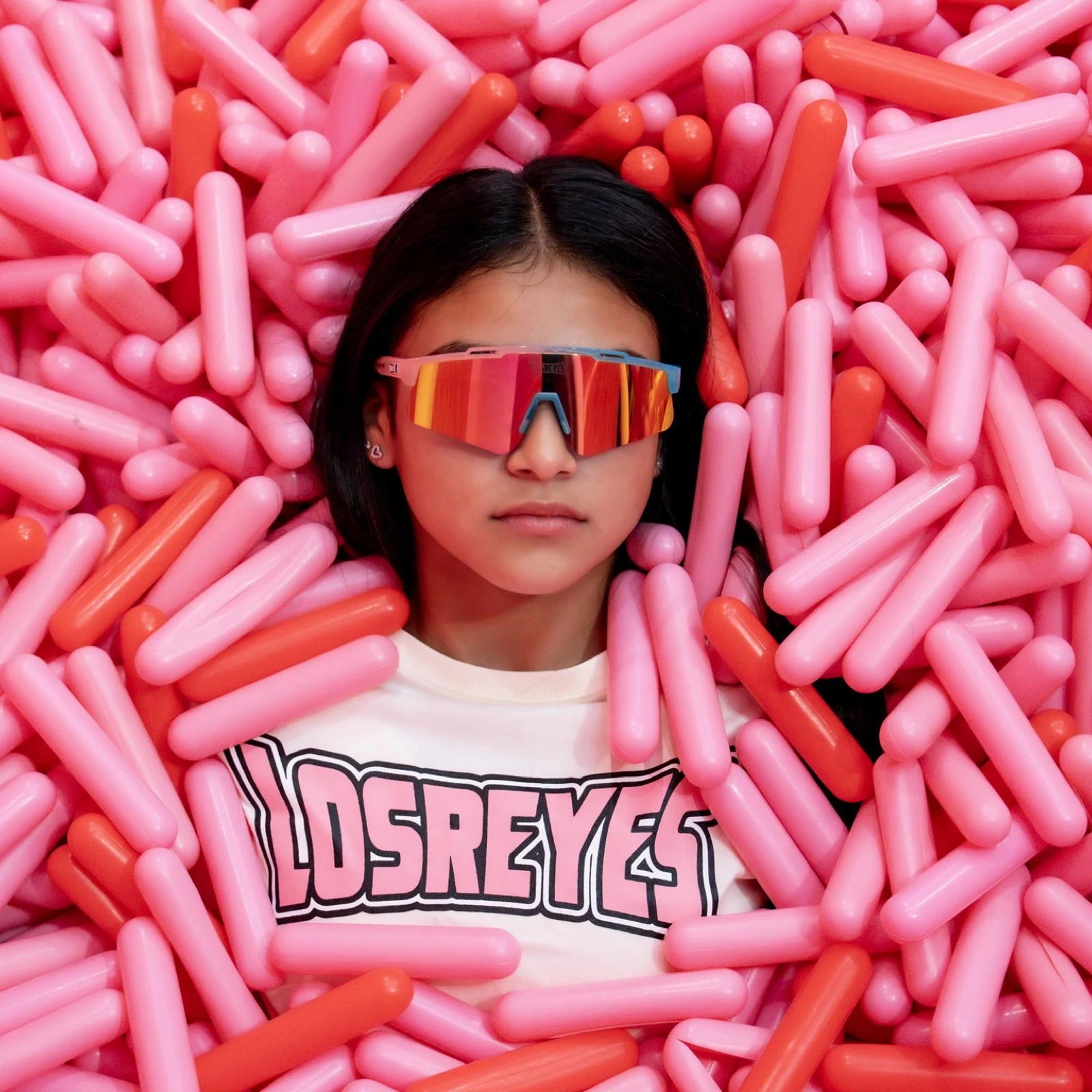 Creative lifestyle image of LosReyes Prime Youth Cotton Candy sports sunglasses worn by girl in pink and red capsule pit, emphasizing mirrored UV400 performance lens and playful, youth-focused athletic style