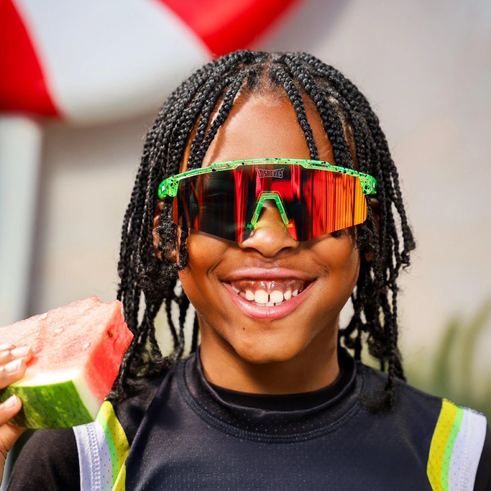 Hero angle of LosReyes Prime Youth Watermelon mirrored shield sunglasses with red-orange lenses, green speckled wraparound frame, and UV400 protection – shown with watermelon for playful summer vibe.