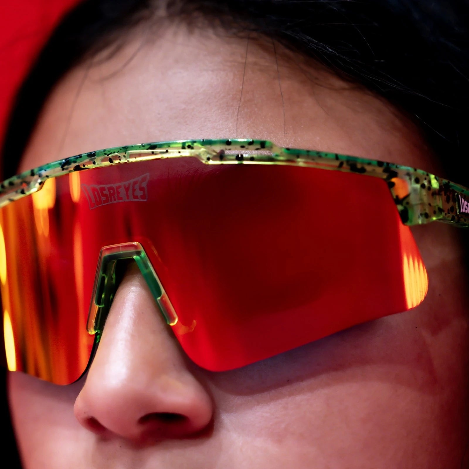 Close-up of LosReyes Prime Youth Watermelon sports sunglasses showcasing orange-red mirrored shield lenses, green splatter frame, and premium UV400 lens clarity.