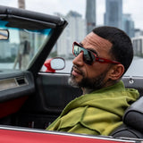 Man wearing LosReyes Ares sunglasses and a green jacket sitting in a convertible car with cityscape in the background
