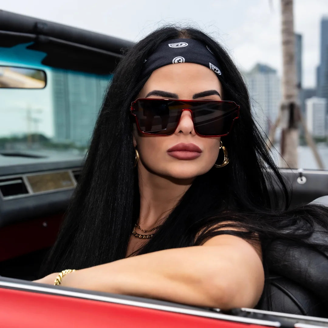 Woman wearing LosReyes sunglasses and a headband, sitting in a convertible car with a cityscape background.